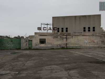 Davanti a noi si presenta il massiccio muro di cinta del complesso, sovrastato dall'insegna recante il nome della societ� e interrotto da un cancello verde sigillato con catene arruginite